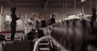 Handheld shot of Muslim sportswoman looking in mirror and doing biceps curls with dumbbells during weightlifting workout in spacious gym