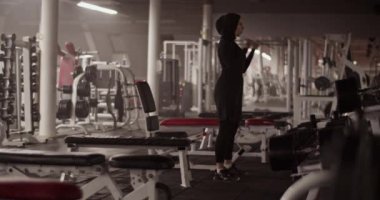 Spor kıyafetleri ve tesettürlü kadın sporcunun düşük açılı el kamerası çekimi ve spor salonunda spor yaparken dambıllarla biceps kıvrımları yapmak.
