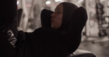 Handheld shot of strong Muslim female athlete in hijab holding hands behind head and doing elevated crunches during fitness workout in gym