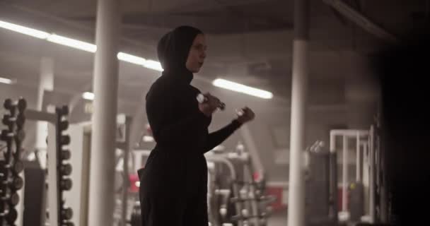 体操锻炼中身穿黑色运动服 头戴头巾 呼吸和用哑铃做二头肌卷的女运动员的低角度手持照片 — 图库视频影像
