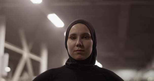 フィットネスワークアウト中にカメラを見てヒジャーブで自信を持ってイスラム教徒のスポーツ女性のハンドヘルドショット照明ジム — ストック動画