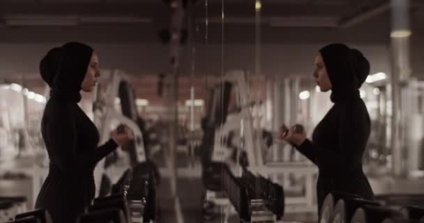イスラム教徒の女性アスリートの手によって撮影された活動的な姿とヒジャーブ鏡を見て ジムでの機能訓練中にダンベルと二足のカールを行う — ストック動画