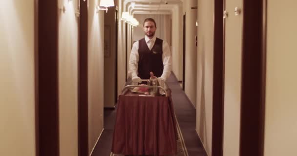 ルームサービス中にホテルの廊下で注文とエレガントな制服ローリングカートでフレンドリーな若い髭の民族のウェイターの完全なボディ — ストック動画