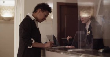Kaliteli giyinmiş kısa kıvırcık saçlı, oteldeki kayıt sırasında resepsiyonistin yanındaki belgeleri imzalayan Afro-Amerikan kadın misafirin yan görüntüsü.