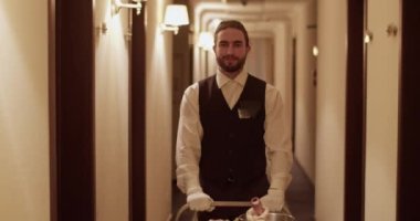 Positive young bearded ethnic room service waiter in elegant uniform smiling and looking at camera while rolling trolley with fruits and wine in hotel hallway