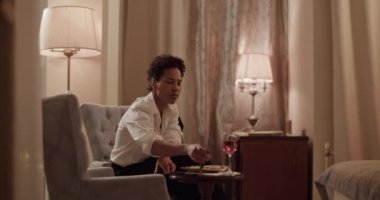 Elegant adult African American businesswoman in classy wear having dinner sitting on comfortable armchair in hotel room during business trip 