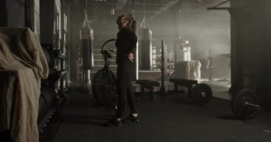 Spor kıyafetleriyle uzun kahverengi saçlı, kaslı genç bayan sporcunun modern ferah spor salonunda yürürken fonksiyonel egzersizler sırasında dinlenmesinin yan görüntüsü.