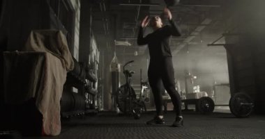 Modern spor salonunda egzersiz yaparken uzun at kuyruğu siyah eşofmanlı, çömelme hareketleri yapan ve omzundan ağır ilaç topu atan güçlü genç bayan sporcu.