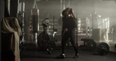 Modern spor salonunda egzersiz yaparken uzun at kuyruğu siyah eşofmanlı, çömelme hareketleri yapan ve omzundan ağır ilaç topu atan güçlü genç bayan sporcu.