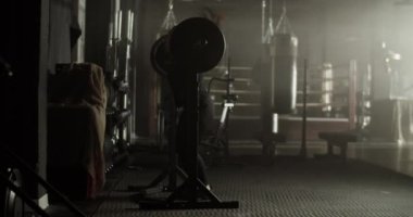 Aktif giyimli, uzun siyah saçlı, ağır halterli, modern spor salonunda egzersiz yaparken çömelmiş güçlü genç bir kadının yan görüntüsü.