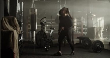 Modern spor salonunda egzersiz yaparken uzun at kuyruğu siyah eşofmanlı, çömelme hareketleri yapan ve omzundan ağır ilaç topu atan güçlü genç bayan sporcu.