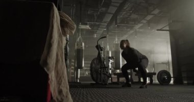 Uzun at kuyruklu güçlü genç, kaslı bayan sporcu spor kulübündeki antrenman sırasında çömelmiş ve kettlebell salıncak egzersizi yapıyor.