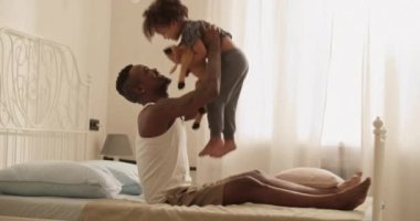 Mutlu, genç, sakallı, sıradan kıyafetleriyle yatakta oturan ve hafta sonu evde oynarken sevimli, mutlu küçük kızını kucaklayan siyah baba.