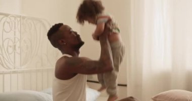 Heyecanlı, genç, sakallı Afrikalı Amerikalı bir babanın günlük giysiler içinde gün ışığında yatakta oturan komik oğluyla oynaşması.