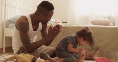 Genç, sakallı Afrikalı Amerikalı baba, yerde oturup, yatak odasında resim yaparken alkışlıyor ve gülümsüyor.