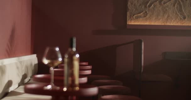 太陽の光の下で現代の公共レストランで小さな丸い赤いテーブルの上に置かれた白ワインボトルとワイングラスのラックフォーカスハンドヘルドショット — ストック動画