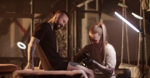 穿着T恤 戴着手套 留着一头长发的年轻女艺人坐在沙龙沙发上 在残忍的大胡子男性顾客腿上纹身的侧面照片 — 图库视频影像