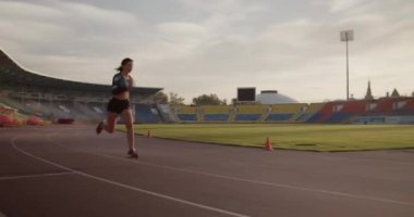 Spor müsabakalarında bulutlu gökyüzüne karşı güneş doğmadan önce stadyumdaki sprint antrenmanı sırasında hızlı koşan genç formda bir bayan koşucunun yan görüş görüntüsü.