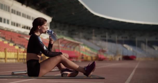 全身而退的年轻疲惫女子短跑运动员全身而退 穿着运动服坐在跑道上 呼吸急促 经过紧张的训练后从瓶子里喝水 — 图库视频影像
