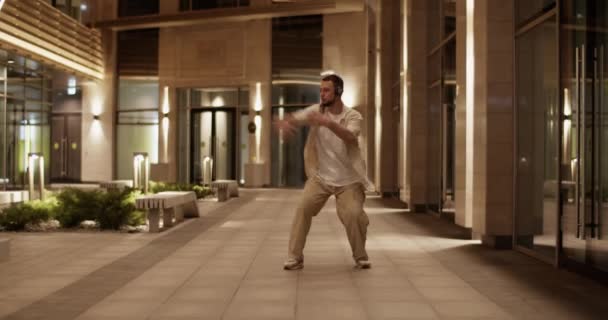 ヘッドフォンダンスで現代の千年男のフルボディリアルタイムショット夜の建物の舗装上のエネルギーでフリースタイル — ストック動画