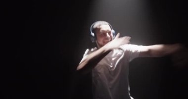 Aktif sakallı erkek dansçı beyaz t-shirt giyip kulaklıkla müzik dinlerken karanlık stüdyoda spot ışıklarıyla break dans yapıyor. 