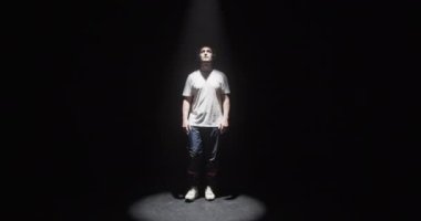 Beyaz tişörtlü ve kulaklıklı beyaz erkek dansçının tüm vücudu karanlığın içinden sahne ışığına çıkıyor sonra sakin müzik dinlerken çağdaş dans ediyor.