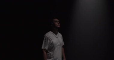 Gerçek zamanlı genç sakallı erkek dansçı karanlık stüdyoda kulaklıkla müzik dinliyor spot ışıklarına bakıyor ve çağdaş dans gösterisi yapıyor.