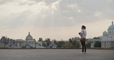 Genç turistin, omuzlarında çantasıyla kaldırımda durup fotoğraf çekerken, bulutlu gökyüzünün altında, donuk güneş ışınları altında inanılmaz şehir manzarasına karşı tüm vücudunu gösteren bir fotoğraf.