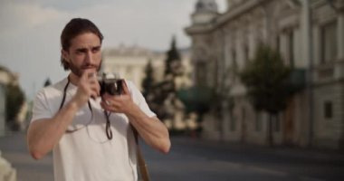 Genç sakallı bir erkek fotoğrafçının eski bir fotoğraf makinesini iterken gerçek zamanlı el kamerası görüntüsü. Sonra da bulanık arka plana doğru yaz yolculuğu sırasında eski şehrin fotoğraflarını çekiyor.