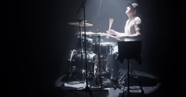 Side view full body of talented young male musician in casual outfit and cap performing song on various drums with sticks on stage in dark studio