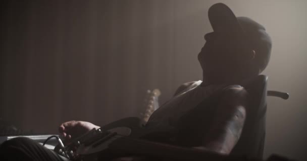 薄暗いライトの録音スタジオで働いている間椅子で振動の帽子の側面のビューのギタリストの静的なショット — ストック動画