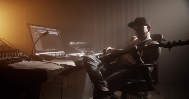 ダークミュージックスタジオでデスクに座ってベースギターを弾くサウンドプロデューサーのハンドヘルドショットサイドビュー — ストック動画