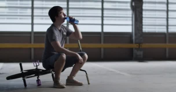 疲れた若い男性Bmxライダーのフルボディハンドヘルドショット 広々とした駐車場で自転車に座って ハードトレーニング後に休んでいる間ボトルから水を飲む — ストック動画