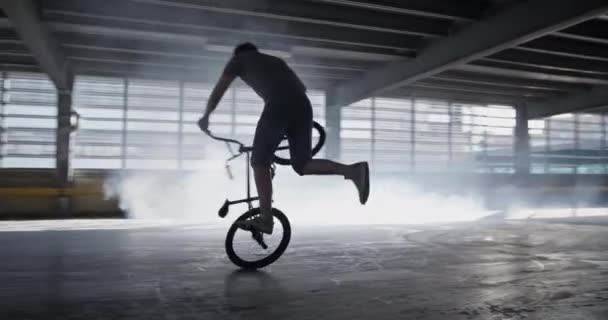 若い男性Bmxライダーのリアルタイム周りパンを自転車で危険なスタントを実行しながら 煙に対する広い駐車場の後ろペグでバランスをとる — ストック動画