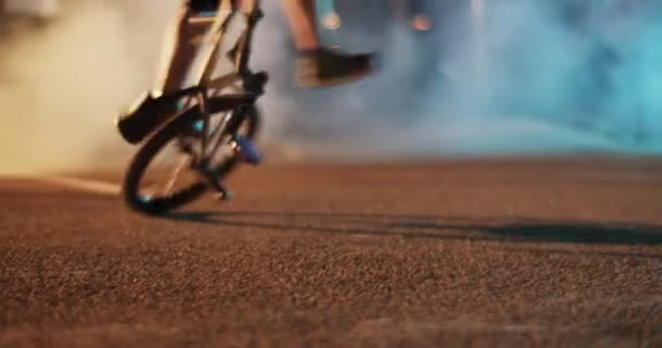 Bmxフラットランドのトリックを実践しながら ネオンライトで照らされた煙に対して舗装された道路上の後輪に自分自身の軸を回転する地上レベルハンドヘルドショット — ストック動画
