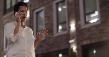 Ofis binasının önünde beyaz gömlekli Afro-Amerikan iş kadınının cep telefonuyla müşterinin iş detaylarını anlattığı düşük açılı el kamerası görüntüsü.