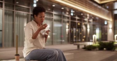 Afro-Amerikan kadın girişimcinin gerçek zamanlı el kamerası görüntüsü. Sıradan giyinmiş, bankta oturmuş, yemek çubuklarıyla erişte yerken ofis binasının dışında dinleniyor.