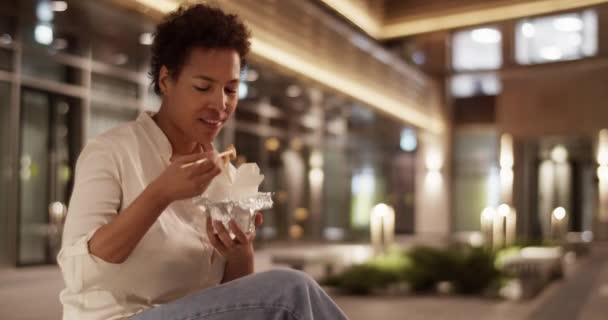 アフリカ系アメリカ人女性起業家のリアルタイムハンドヘルドショットは ベンチに座って 外のオフィスビルで休憩しながら箸で麺を食べるオーダーメイドを楽しんでいます — ストック動画