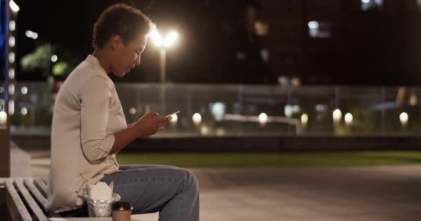 サイドビューアフリカ系アメリカ人のビジネスマンの笑顔のリアルタイム ベンチに座ってスマートなカジュアルな服を注文された食べ物と仕事後の夜の都市で携帯電話を介して友人を呼び出す — ストック動画
