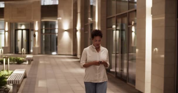 アフリカ系アメリカ人大人のビジネスマンの追跡ショット 夜間のオフィスビルの外を歩く短いアフリカの髪と携帯電話の完成作業を介してメッセージング — ストック動画