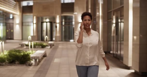 アフリカ系アメリカ人女性ディレクターのトラッキングショットは 携帯電話で話しながら輝くランプでビジネスセンターの外を歩き 夜の時間に仕事の問題を議論 — ストック動画