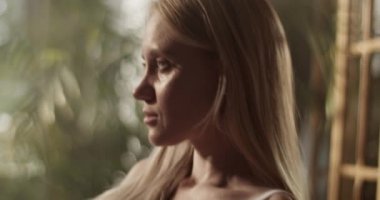 Resim stüdyosunda beyaz elbiseli genç bir kadın ressamın güneş ışığıyla aydınlatılmış ve resim yaparken elinde fırçayla oturduğu el kamerasıyla çekilmiş bir fotoğraf.