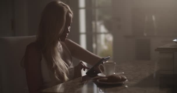スマートフォン経由でソーシャルメディアをスクロールしながら薄暗いキッチンでホットドリンクのマグカップとテーブルに座っている若い女性のサイドビューハンドヘルドショット — ストック動画