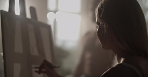 薄暗い光で照らされたスタジオに座る女性画家のソフトフォーカスリアルタイムと ライフデッサンセッション中にコットンキャンバスに鉛筆で男性モデルからスケッチを描画 — ストック動画