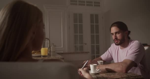 テーブルに座って長髪の若い男性モデルのリアルタイムハンドヘルドショット キッチンでアイデアを共有しながら画家と将来の絵画を議論 — ストック動画