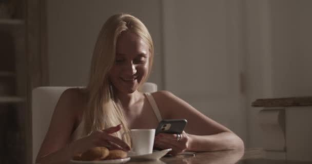 キッチンサーフィンソーシャルメディアで携帯電話を閲覧しながら美味しいホットドリンクを楽しんでテーブルに座っている若いポジティブなブロンドの女性のリアルタイムハンドヘルドショット — ストック動画