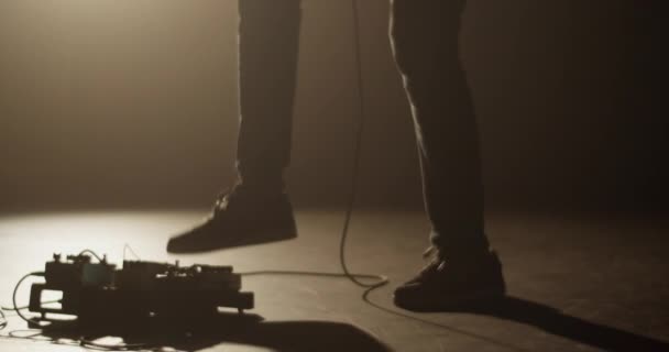 イエロースポットライトで照らされたダークスタジオで音楽を演奏しながら 足で電気ギターのペダルスタックを押す匿名の男性ミュージシャンのハンドヘルドショットの周りパン — ストック動画