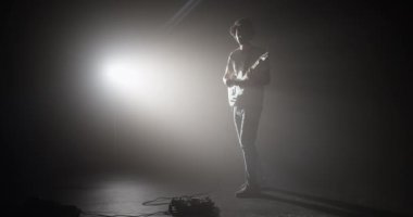 Karanlık stüdyoda durup müzik aletinden yansıyan loş spot ışıklarıyla müzik enstrümanı çalan etkileyici erkek gitaristin tam vücut resmi.