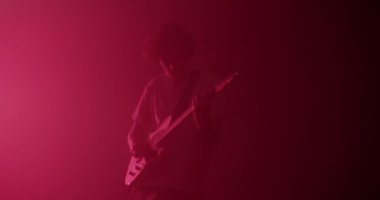 Karanlık stüdyoda pembe neon ışıkla hızlı gitar çalarken hareketli ve titreyen dışavurumcu erkek gitarist.