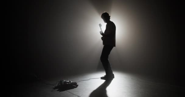 パフォーマンス中に輝く光と電気楽器を演奏するために暗闇に立っているエネルギッシュな男性ギタープレーヤーのリアルタイムフルボディ — ストック動画